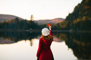 Rückenansicht einer nicht erkennbaren jungen stilvollen Touristin in elegantem rotem Mantel und weißer Baskenmütze, die ein Ahornblatt zeigt, während sie am Ufer eines ruhigen Teiches inmitten eines üppigen Herbstwaldes steht - ADSF35625