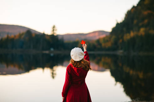 Back view of unrecognizable young stylish female tourist in elegant red coat and white beret showing maple leaf while standing on shore of calm pond surrounded by lush autumn forest - ADSF35625