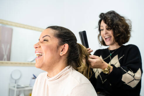 Cheerful brunette using iron to curl hair of glad female client during work in beauty salon - ADSF35631