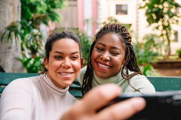 Cheerful black woman in sweater laughing and taking selfie with Hispanic girlfriend while sitting on bench in park - ADSF35633