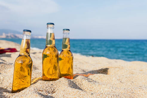 Glasflaschen mit kaltem Bier auf Sand auf unscharfen Hintergrund von Meer und blauem Himmel auf sonnigen Tag auf Resort platziert - ADSF35638