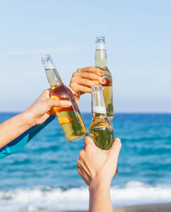 Anonymer Ausschnitt der Hände eines Mannes und einer Frau, die eine Bierflasche hochhalten, während sie am Strand gegen das plätschernde Meerwasser und den blauen Himmel an einem Sommerwochenende chillen - ADSF35643