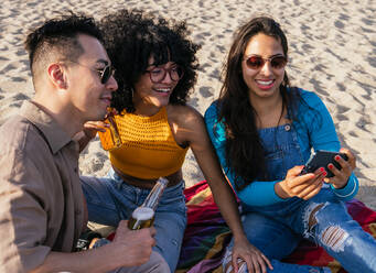 Happy multiracial man and women smiling and browsing social media on cellphones while resting on beach on sunny summer day on resort - ADSF35644