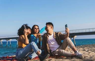 Mann und Frauen in Freizeitkleidung mit Bier sitzen auf Sandstrand und nehmen Selfie über Smartphone gegen Meerwasser und blauen Himmel - ADSF35645