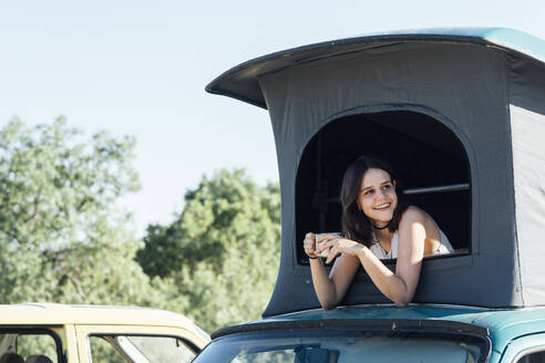 Happy young woman with coffee cup looking through roof tent - DAMF01020