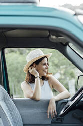 Happy young woman wearing hat leaning on van window - DAMF01028