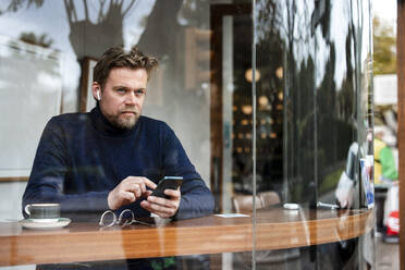 Älterer Geschäftsmann mit Mobiltelefon in einem Café sitzend, gesehen durch ein Glasfenster - JOSEF11035