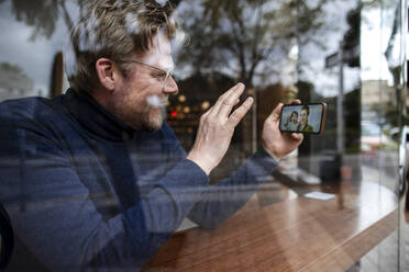Glücklicher Geschäftsmann winkt seiner Familie bei einem Videoanruf über ein Smartphone im Café zu - JOSEF11053