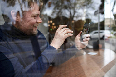 Glücklicher Geschäftsmann im Gespräch mit seiner Tochter bei einem Videoanruf in einem Café, gesehen durch ein Glasfenster - JOSEF11056