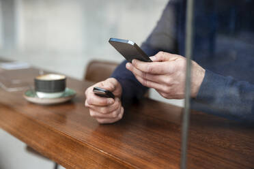 Hände eines Geschäftsmannes, der einen Autoschlüssel hält und ein Smartphone in einem Café benutzt, gesehen durch ein Glasfenster - JOSEF11066