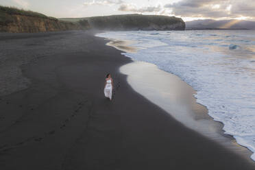 Luftaufnahme einer Frau in einem weißen Kleid am Strand mit Klippen im Hintergrund, Sao Miguel, Azoren, Portugal. - AAEF14941