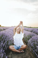 Smiling woman with arms raised sitting amidst lavender field - SIF00343