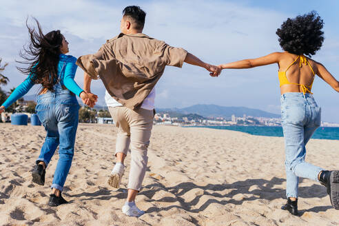 Rückansicht eines aufgeregten multiethnischen Mannes und einer Frau, die sich an den Händen halten und auf dem Sand gegen den blauen Himmel laufen, während sie einen Sommertag in der Nähe des Meeres am Urlaubsort verbringen - ADSF35646