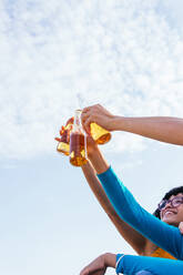 Von unten anonym abgeschnittene Hände eines Mannes und einer Frau, die Bierflaschen hochhalten, während sie am Strand gegen den blauen Himmel an einem Sommerwochenende chillen - ADSF35648