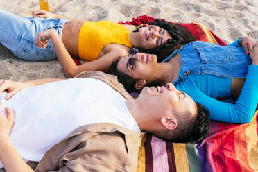 Diverse Männer und Frauen in Freizeitkleidung liegen auf einer Decke und lächeln, während sie Zeit am Sandstrand in der Nähe des Meeres an einem Sommerwochenende verbringen - ADSF35651