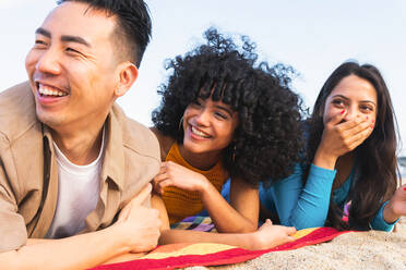 Diverse Männer und Frauen in Freizeitkleidung liegen auf einer Decke und lächeln, während sie Zeit am Sandstrand in der Nähe des Meeres an einem Sommerwochenende verbringen - ADSF35654