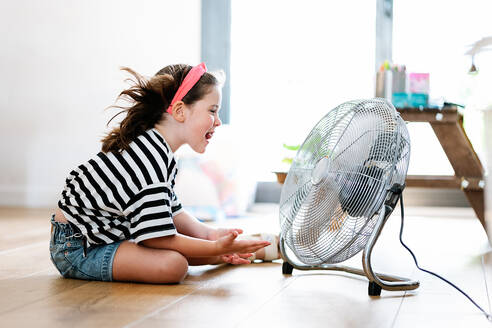 Seitenansicht eines kleinen Mädchens, das zu Hause auf dem Boden sitzt und mit einem Ventilator spielt - ADSF35656