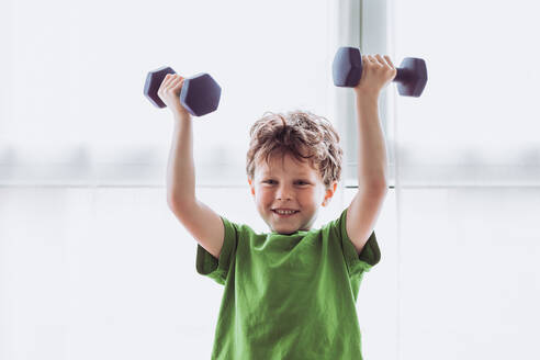 Fröhliches Kind in Sportkleidung, das lächelt und mit Hanteln gegen das Fenster beim Fitnesstraining am Morgen zu Hause trainiert - ADSF35665