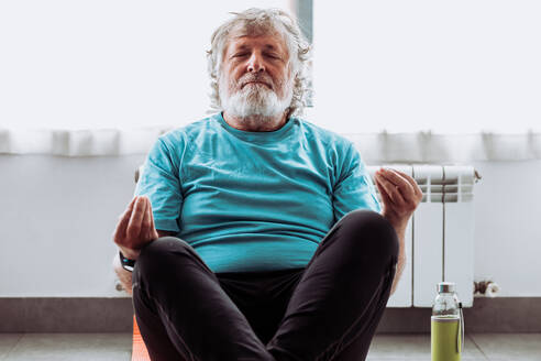 Aged male in activewear sitting on floor in Lotus pose and meditating with closed eyes during yoga session in morning at home - ADSF35670