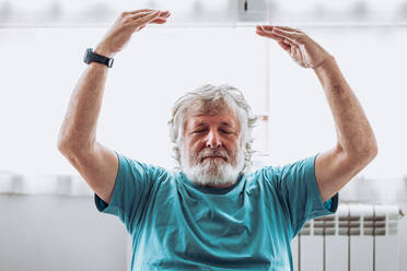 Älterer Mann in blauem T-Shirt hebt die Arme und meditiert mit geschlossenen Augen gegen das Fenster während einer Yoga-Sitzung am Morgen zu Hause - ADSF35671