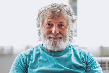 Älterer Mann mit grauem Bart vor unscharfem Hintergrund schaut zu Hause in die Kamera - ADSF35672