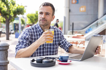Älterer digitaler Nomade sitzt mit Laptop und trinkt Orangensaft in einem Straßencafé - WPEF06165