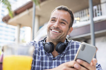 Happy mature man with smart phone and wireless headphones at cafe - WPEF06171