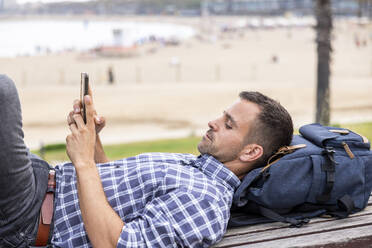 Älterer Mann liegt auf einer Bank und benutzt ein Mobiltelefon am Strand - WPEF06181