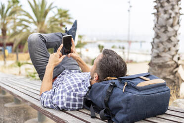 Älterer Mann benutzt sein Smartphone und entspannt sich auf einer Bank am Strand - WPEF06183