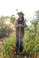 Full body little gardener looking at camera in casual clothes using hoe to remove weeds from soil near green scallions while working in sunlit field in summer - ADSF35696