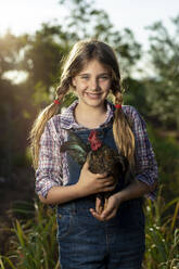 Positives Mädchen in Freizeitkleidung mit Hahn, der lächelt und in die Kamera schaut, während er einen sonnigen Sommertag auf dem Bauernhof verbringt - ADSF35698