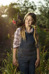 Positives Mädchen in Freizeitkleidung mit Hahn lächelnd bei sonnigen Sommertag auf dem Bauernhof - ADSF35699