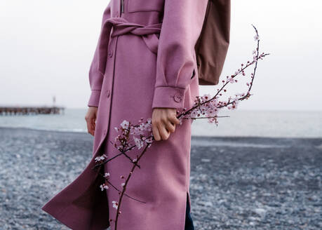 Cropped unrecognizable female in pink coat standing with blooming cheery flower twig - ADSF35702