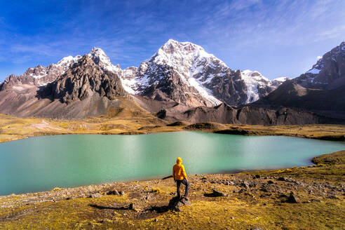 Tourist in yellow hoodie standing on coast of calm turquoise lake and admiring snowy peaks of Ausangate mountain on sunny day in Peru - ADSF35703