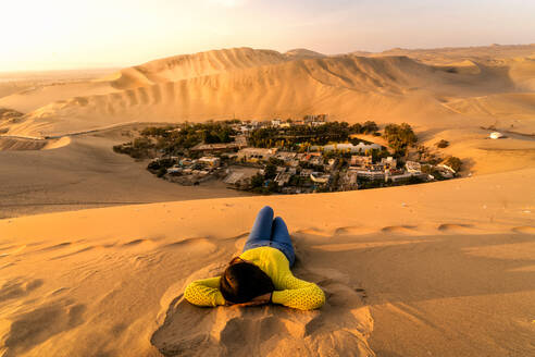 Unrecognizable female traveler with dark hair in casual outfit lying on sand dunes and admiring picturesque Huacachina village against sunset sky in Peru - ADSF35706