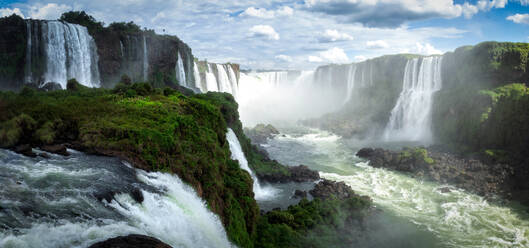 Atemberaubende Landschaft der mächtigen Iguazu-Wasserfälle, die durch felsige, mit grünen Pflanzen bedeckte Klippen fließen, vor einem bewölkten blauen Himmel an der Grenze zwischen Argentinien und Brasilien - ADSF35707