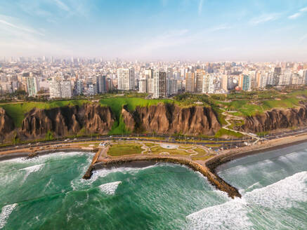 Breathtaking aerial view of wavy ocean washing sandy coastline near modern residential and commercial building against cloudy blue sky in Lima - ADSF35713