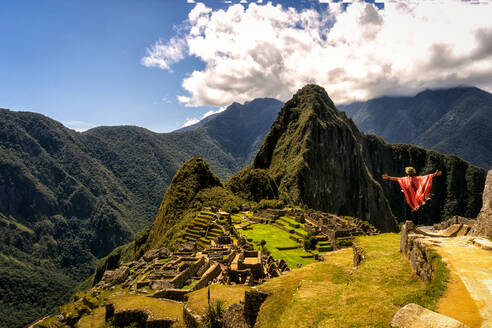 Back view of unrecognizable traveler in traditional poncho and hat admiring scenic mountainous valley while standing on viewpoint with outstretched arms during trip in Machu Picchu - ADSF35717