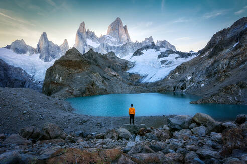 Rückenansicht eines nicht erkennbaren Touristen in gelber Jacke und Hut, der einen malerischen See bewundert, der von massiven felsigen Bergen mit schneebedeckten Hängen umgeben ist, während einer Reise in der Region Patagonien in Argentinien - ADSF35721