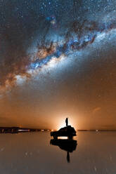 Silhouette eines nicht erkennbaren Reisenden, der auf einem Geländewagen steht, der auf der glänzenden Oberfläche der Salzwüste von Salar De Uyuni geparkt ist, und der die am Abendhimmel leuchtende Milchstraße bewundert - ADSF35725