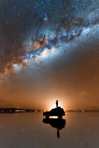 Silhouette of unrecognizable traveler standing on SUV car parked on shiny surface of salt flat of Salar De Uyuni and admiring scenic Milky Way galaxy shining in evening sky - ADSF35725