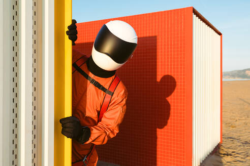 Anonymer Kosmonaut in orangefarbenem Raumanzug und futuristischem Helm, der bei der Erkundung einer Basis hinter einer Wand hervorschaut - ADSF35732
