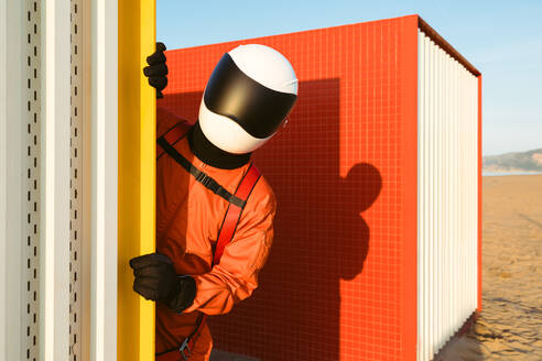 Anonymous cosmonaut in orange spacesuit and futuristic helmet peeking from behind wall while exploring base - ADSF35732