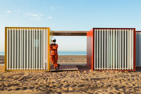 Back view of astronaut in spacesuit and helmet standing with arms crossed on sand near futuristic building and admiring sea against blue sky on alien planet - ADSF35733