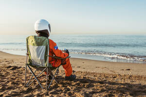 Unbekannter Astronaut in futuristischem Raumanzug und Helm, der auf Sand sitzt und das wogende Meer bewundert, während er abends am Strand verweilt - ADSF35736