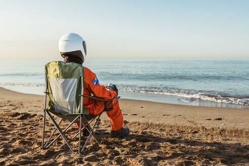 Unrecognizable astronaut in futuristic spacesuit and helmet sitting on sand and admiring waving sea while spending time on beach in evening - ADSF35736