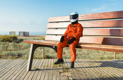 Full body astronaut in sci fi spacesuit and helmet sitting on large bench against blue sky on beach of alien planet - ADSF35737