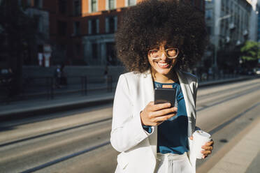 Lächelnde Geschäftsfrau mit Smartphone und Kaffeetasse beim Überqueren der Straße an einem sonnigen Tag - MEUF06898