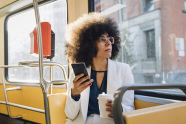 Junge Geschäftsfrau mit Smartphone und Kaffeetasse beim Pendeln im Bus - MEUF06899