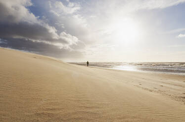 Clouds over sandy beach at sunset with silhouette of man walking in background - GWF07507